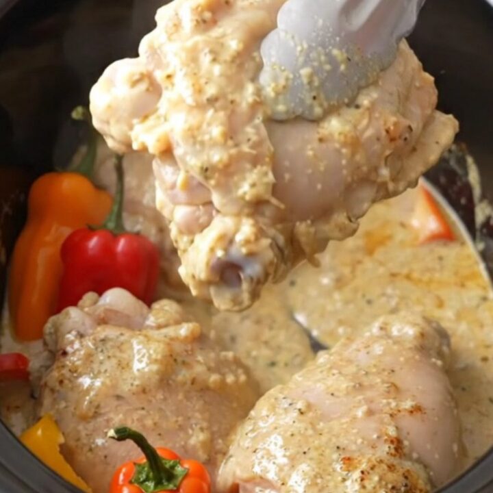 A white slow cooker filled with creamy chicken thighs and vibrant red, orange, and yellow mini peppers.