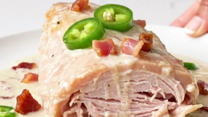 A close-up of a cooked turkey tenderloin topped with creamy sauce, bacon bits, and jalapeno rings on a white plate.