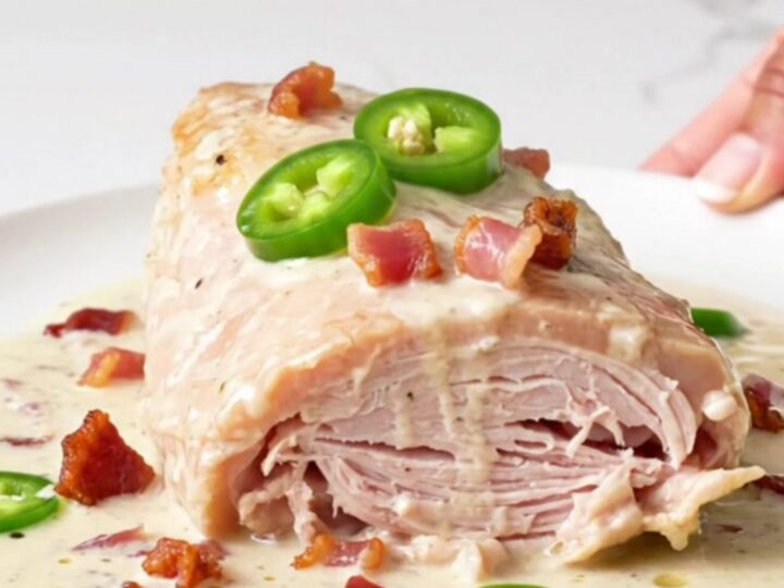 A close-up of a cooked turkey tenderloin topped with creamy sauce, bacon bits, and jalapeno rings on a white plate.