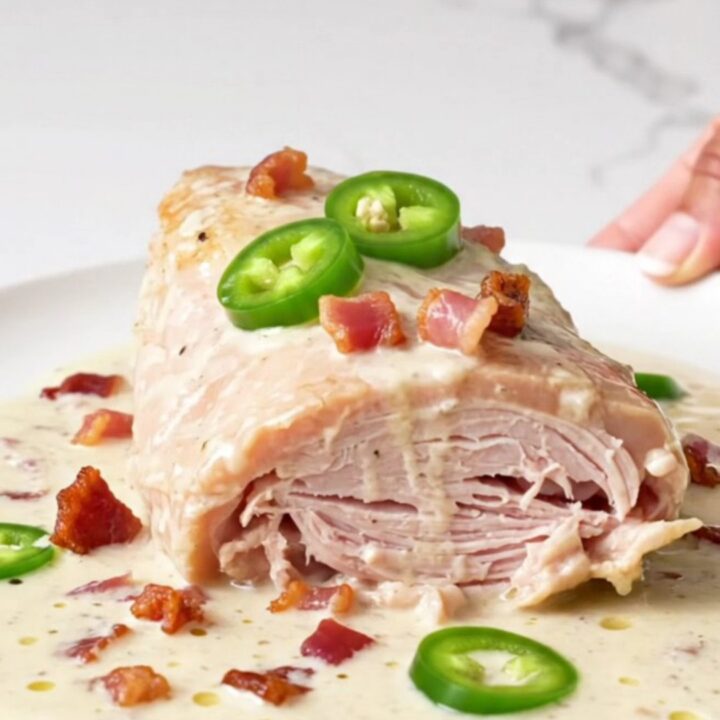 A close-up of a cooked turkey tenderloin topped with creamy sauce, bacon bits, and jalapeno rings on a white plate.