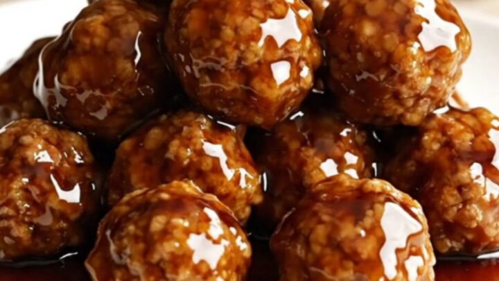 A close-up of honey balsamic meatballs on a white plate, coated in a glossy dark sauce with light steam rising.