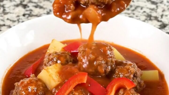 A close-up of a bowl of slow-cooked meatball goulash with tender potatoes and red bell peppers in a rich red sauce.