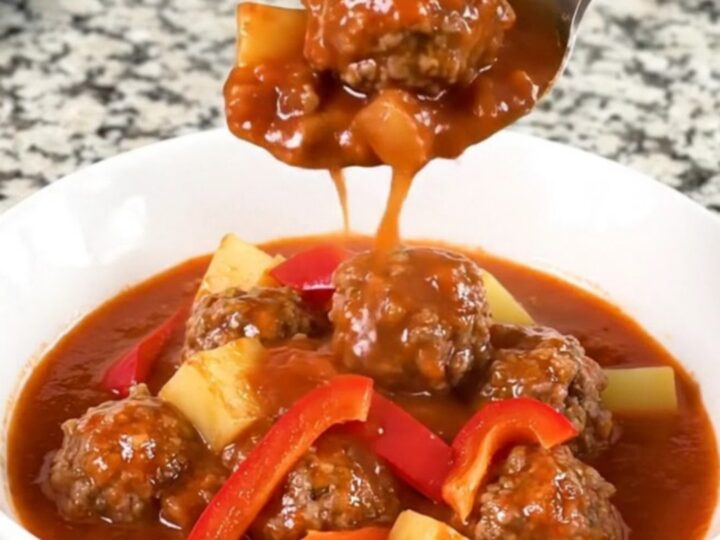 A close-up of a bowl of slow-cooked meatball goulash with tender potatoes and red bell peppers in a rich red sauce.