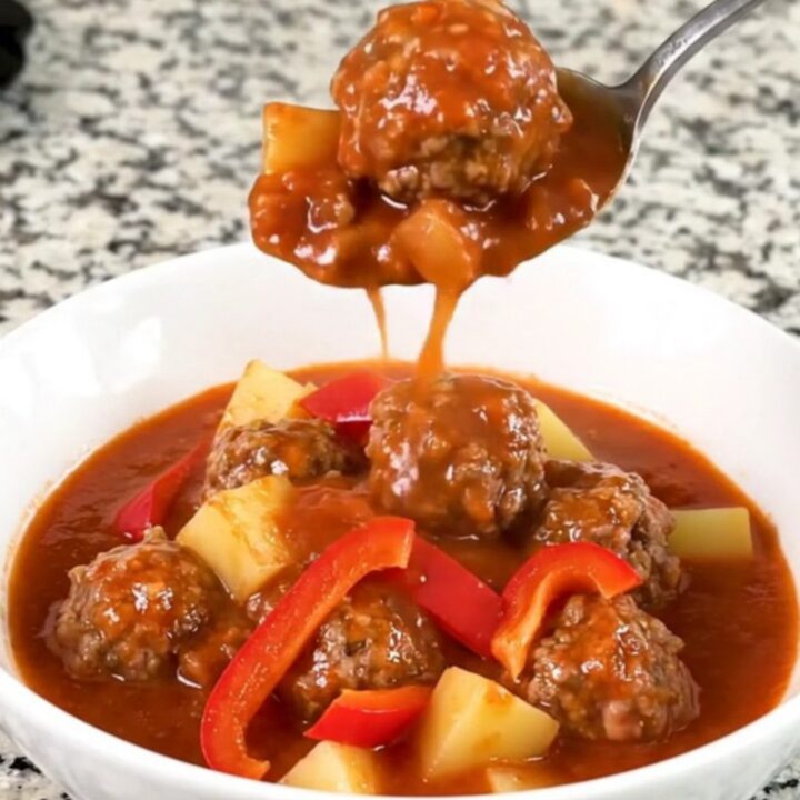 A close-up of a bowl of slow-cooked meatball goulash with tender potatoes and red bell peppers in a rich red sauce.