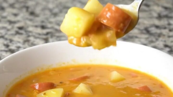 A steaming bowl of creamy orange cheese soup filled with coin-shaped hot dog slices and diced potatoes.