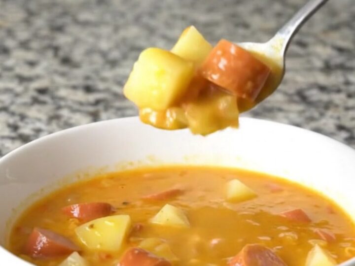 A steaming bowl of creamy orange cheese soup filled with coin-shaped hot dog slices and diced potatoes.
