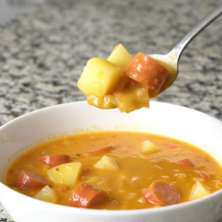 A steaming bowl of creamy orange cheese soup filled with coin-shaped hot dog slices and diced potatoes.