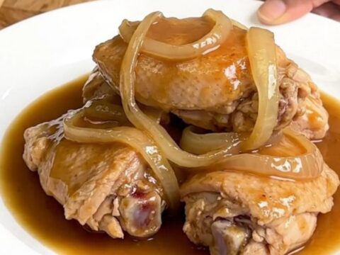 A close-up of tender, braised chicken thighs served with soft, translucent onion slices in a rich brown au jus gravy.