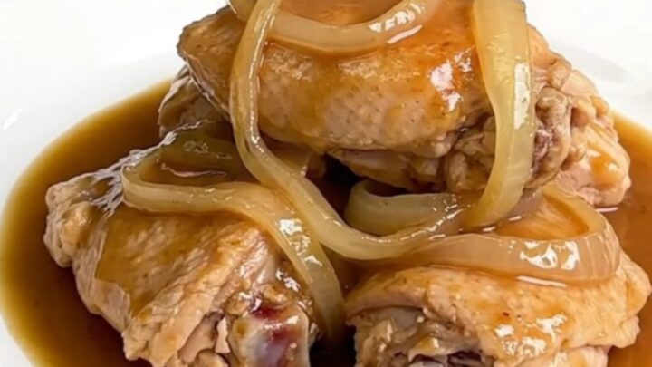 A close-up of tender, braised chicken thighs served with soft, translucent onion slices in a rich brown au jus gravy.