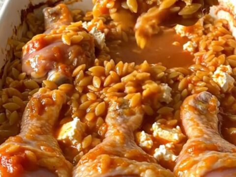 A top-down view of a rectangular baking dish filled with roasted chicken drumsticks and tomato orzo topped with feta.