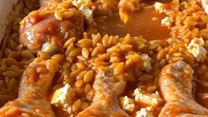 A top-down view of a rectangular baking dish filled with roasted chicken drumsticks and tomato orzo topped with feta.
