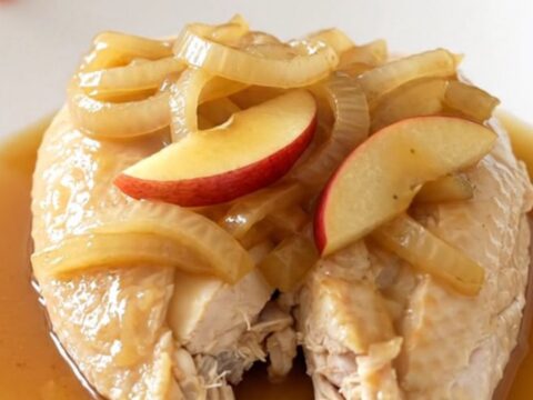 A top-down view of raw chicken breasts, sliced apples, and onions inside a black slow cooker insert.
