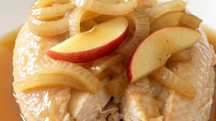 A top-down view of raw chicken breasts, sliced apples, and onions inside a black slow cooker insert.