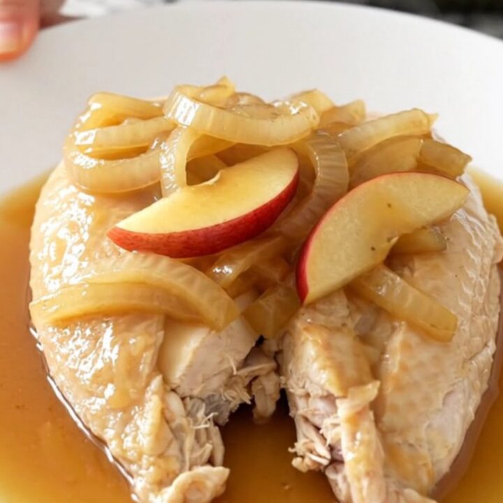A top-down view of raw chicken breasts, sliced apples, and onions inside a black slow cooker insert.