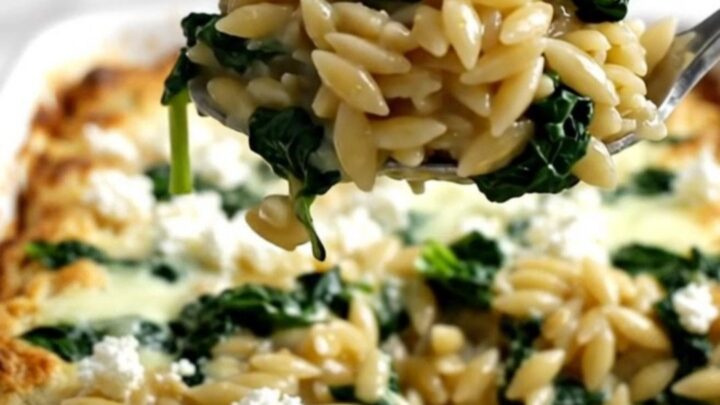 A rectangular baking dish filled with a golden, baked orzo dish topped with crumbled white feta and wilted green spinach.