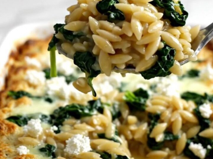 A rectangular baking dish filled with a golden, baked orzo dish topped with crumbled white feta and wilted green spinach.