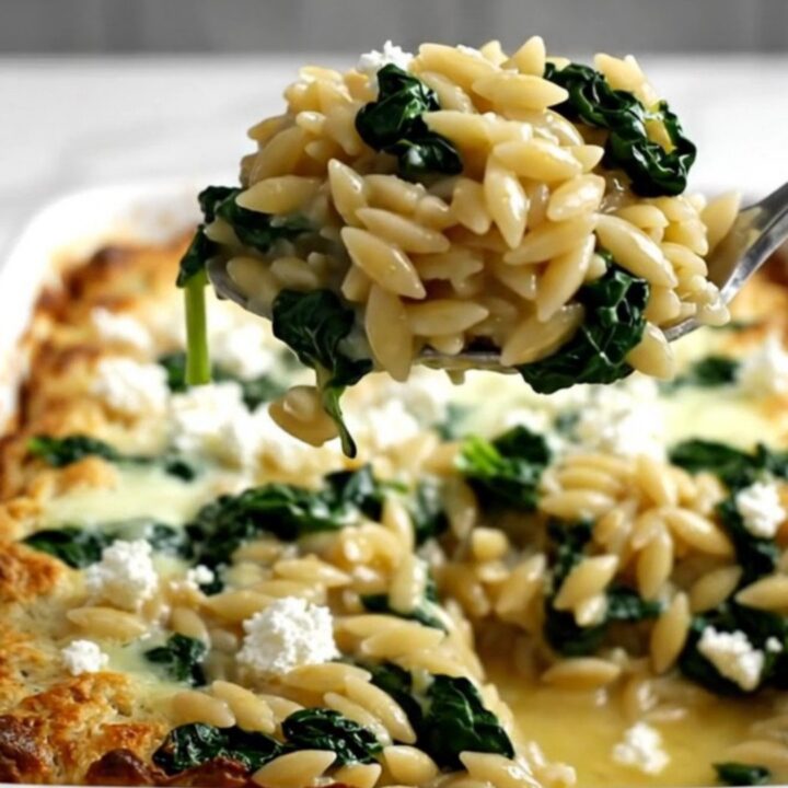 A rectangular baking dish filled with a golden, baked orzo dish topped with crumbled white feta and wilted green spinach.