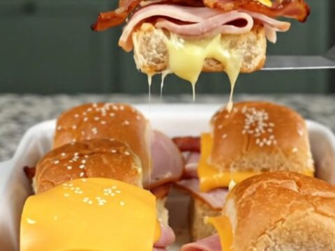 A golden, baked tray of club sandwich sliders on a granite countertop, showcasing melted cheese and sesame-topped buns.