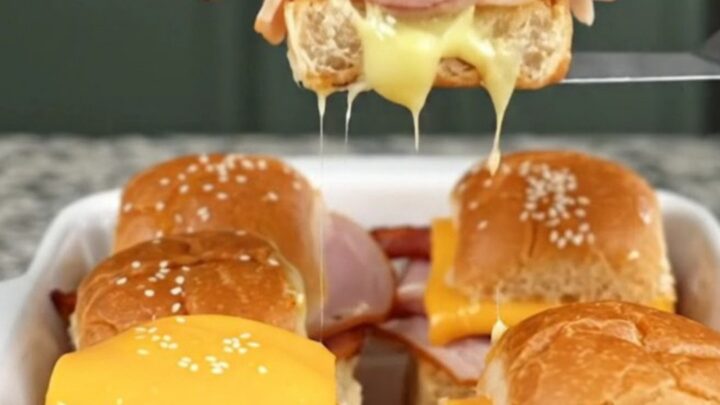 A golden, baked tray of club sandwich sliders on a granite countertop, showcasing melted cheese and sesame-topped buns.