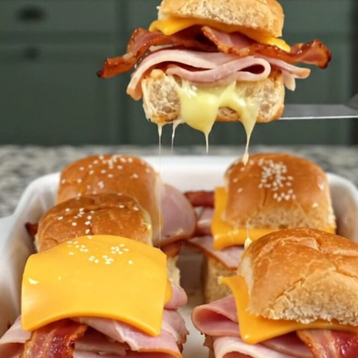 A golden, baked tray of club sandwich sliders on a granite countertop, showcasing melted cheese and sesame-topped buns.