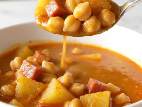 A top-down view of a slow cooker filled with ham, chickpeas, potatoes, and a rich, red paprika-infused broth.