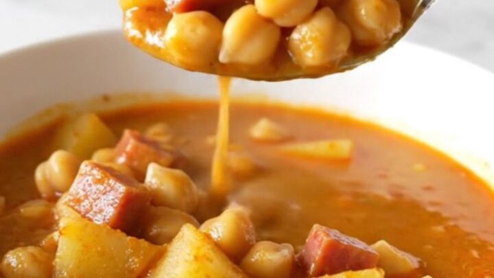 A top-down view of a slow cooker filled with ham, chickpeas, potatoes, and a rich, red paprika-infused broth.
