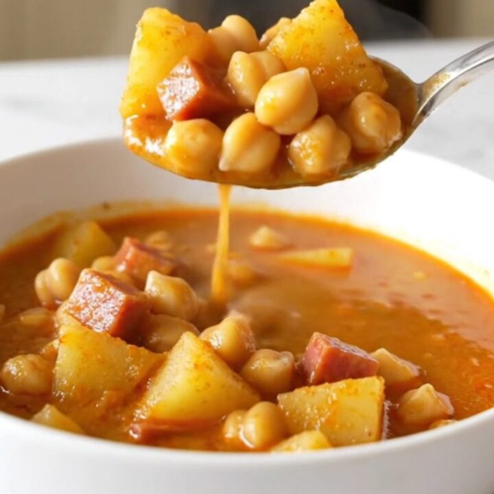 A top-down view of a slow cooker filled with ham, chickpeas, potatoes, and a rich, red paprika-infused broth.