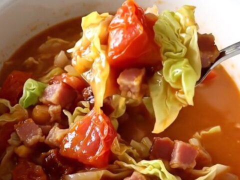 A white bowl filled with slow-cooked cabbage, bacon, and stewed tomatoes, featuring a spoon lifting a bite.
