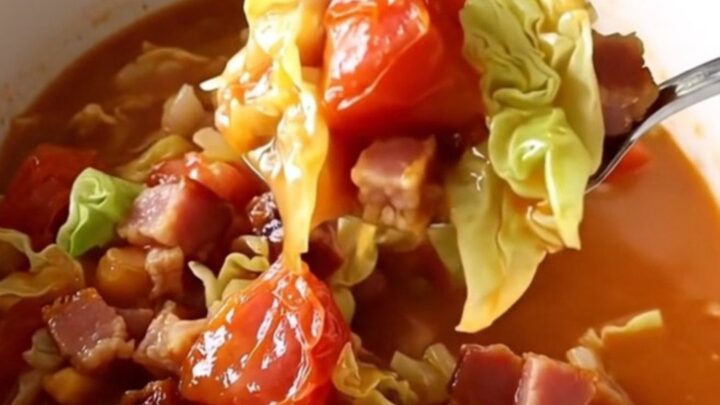 A white bowl filled with slow-cooked cabbage, bacon, and stewed tomatoes, featuring a spoon lifting a bite.