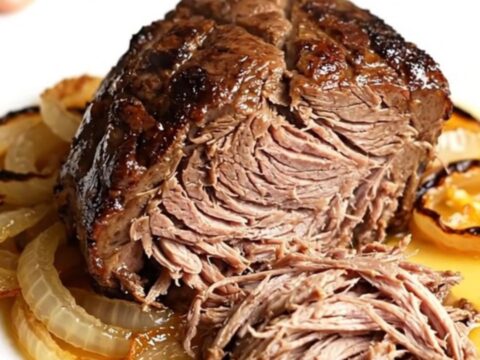 A high-angle shot of a raw beef chuck roast in a black slow cooker insert on a beige countertop.
