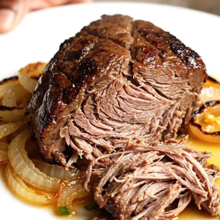 A high-angle shot of a raw beef chuck roast in a black slow cooker insert on a beige countertop.