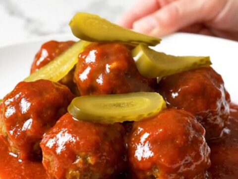 A slow cooker filled with golden chicken meatballs coated in a glossy, vibrant red Nashville hot sauce and dill pickle chips.