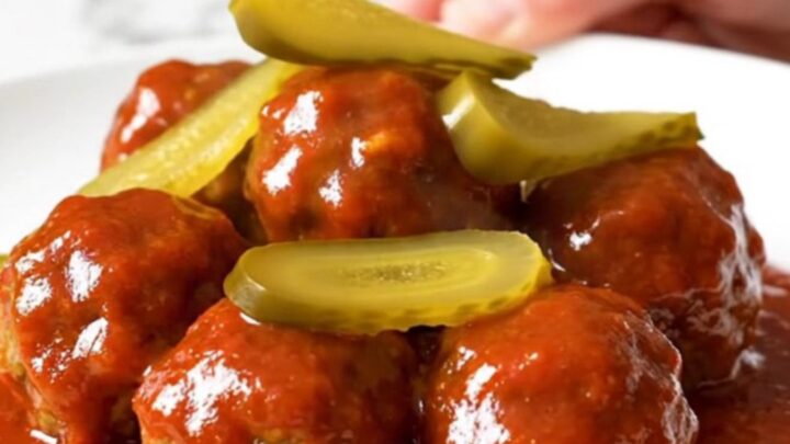 A slow cooker filled with golden chicken meatballs coated in a glossy, vibrant red Nashville hot sauce and dill pickle chips.