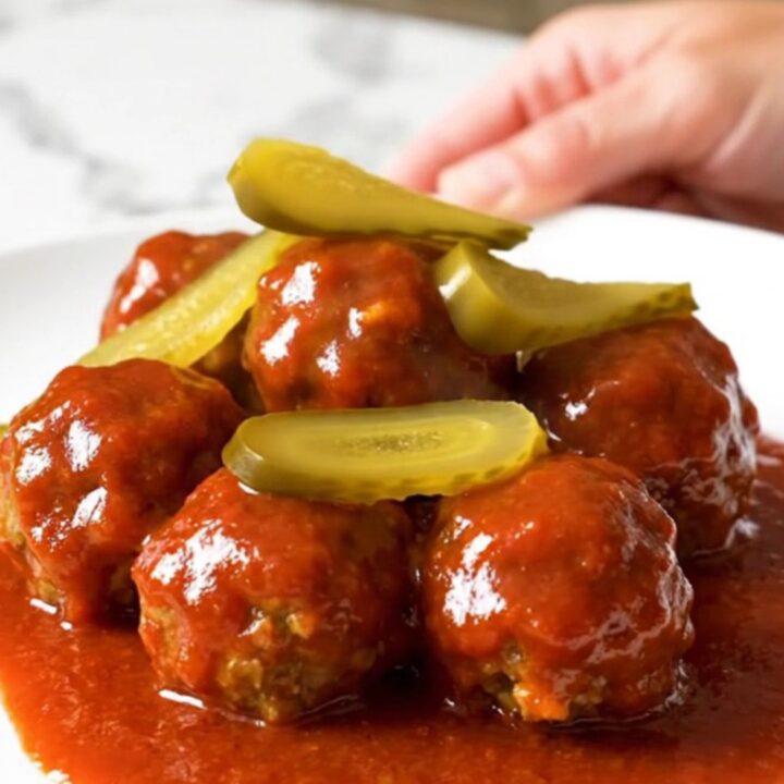 A slow cooker filled with golden chicken meatballs coated in a glossy, vibrant red Nashville hot sauce and dill pickle chips.