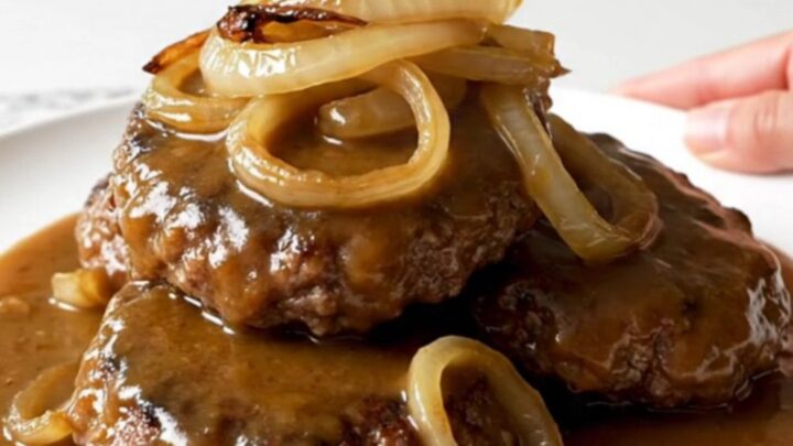 A close-up of tender hamburger steaks covered in caramelized onions and rich brown gravy in a slow cooker.