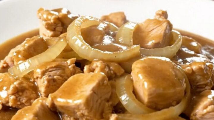 A close-up of tender bite-sized chicken cubes smothered in a rich brown gravy with onion slices in a slow cooker.