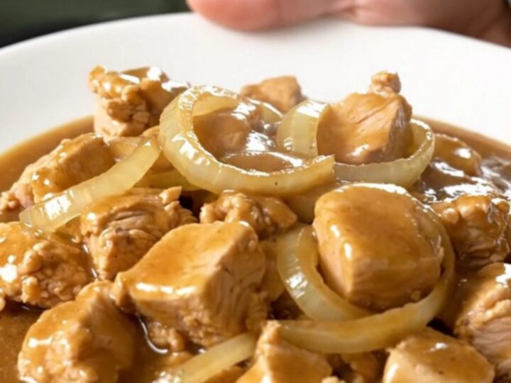 A close-up of tender bite-sized chicken cubes smothered in a rich brown gravy with onion slices in a slow cooker.