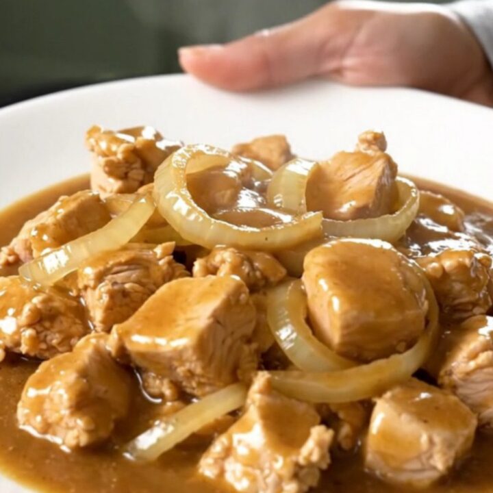 A close-up of tender bite-sized chicken cubes smothered in a rich brown gravy with onion slices in a slow cooker.