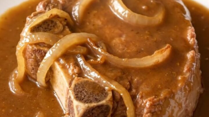 A close-up of tender, slow-cooked bone-in pork steaks covered in a thick brown gravy and sliced onions.