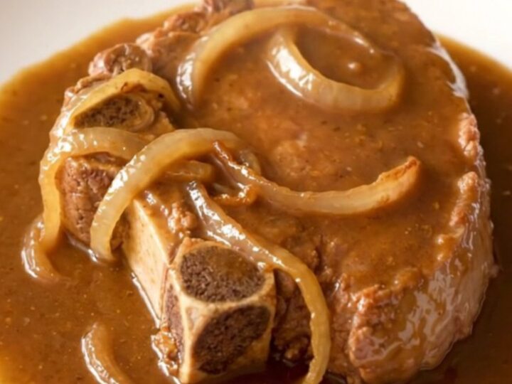 A close-up of tender, slow-cooked bone-in pork steaks covered in a thick brown gravy and sliced onions.
