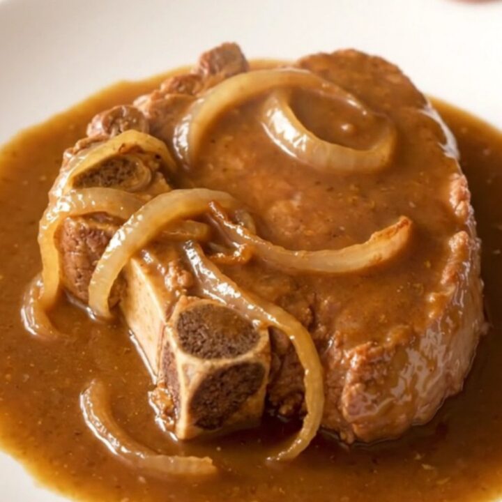 A close-up of tender, slow-cooked bone-in pork steaks covered in a thick brown gravy and sliced onions.