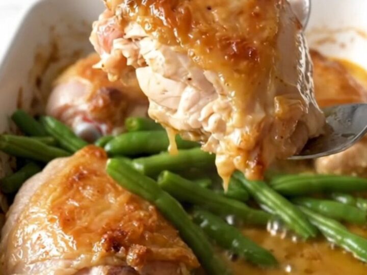 A ceramic baking dish filled with golden baked chicken thighs and bright green beans in a lemon butter sauce.