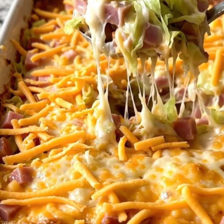Top-down view of a ceramic baking dish filled with fresh cabbage, cubed ham, and creamy sauce topped with shredded cheddar.
