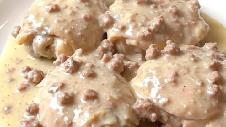 A plate of tender chicken thighs smothered in a thick, creamy sausage gravy with visible herbs.