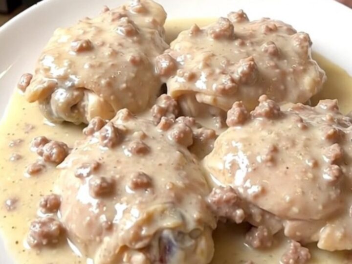 A plate of tender chicken thighs smothered in a thick, creamy sausage gravy with visible herbs.