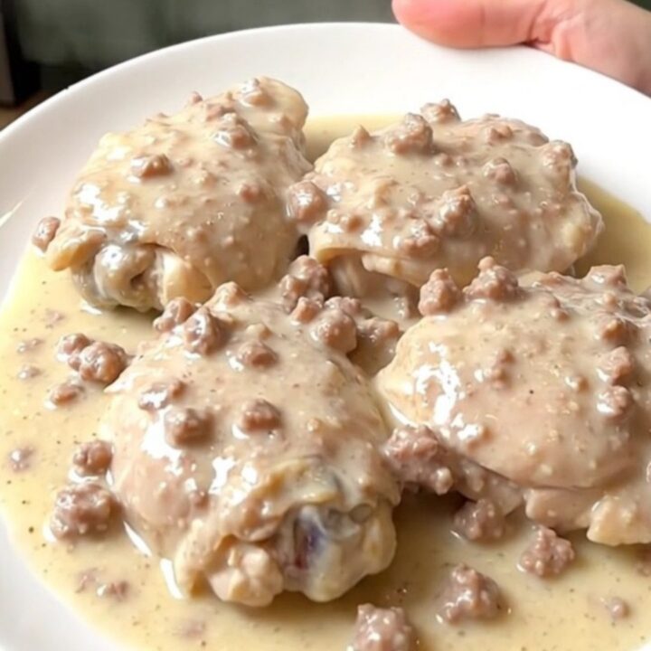 A plate of tender chicken thighs smothered in a thick, creamy sausage gravy with visible herbs.