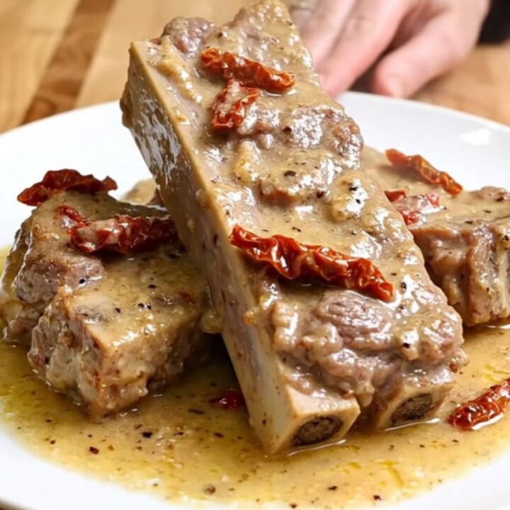 A close-up of tender, creamy pork ribs served on a white plate with sun-dried tomatoes and parmesan sauce.