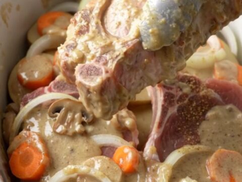 A close-up of a slow cooker filled with tender bone-in pork chops, sliced potatoes, and carrots in a creamy mushroom sauce.