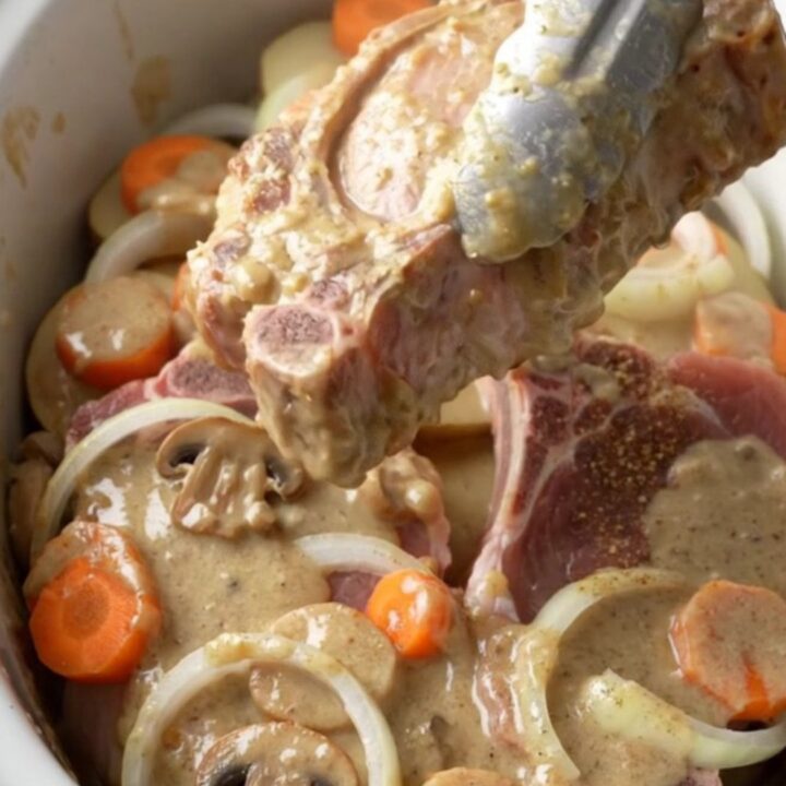 A close-up of a slow cooker filled with tender bone-in pork chops, sliced potatoes, and carrots in a creamy mushroom sauce.