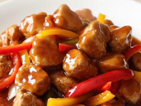 A close-up of tender Beijing chicken pieces coated in a glossy red sauce with bell peppers on a white plate.